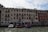 A canal-side scene featuring historic Venetian architecture. The façade of a multi-story building displays warm colors with multiple windows, balconies, and flags. In the canal, two boats are docked near striped mooring poles, and several people are gathered on the shore, possibly near an entrance to the building under an awning.