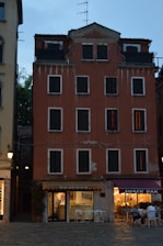 A multi-story building with a reddish facade and dark, shuttered windows. The structure appears worn, with visible peeling paint and signs of aging. The ground floor features a hotel entrance on the left, marked 'Hotel San Geremia,' and a snack bar on the right with people seated outside. The scene is set in the early evening with artificial lights illuminating the snack bar area.
