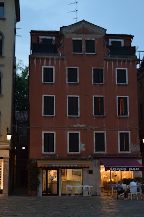 A multi-story building with a reddish facade and dark, shuttered windows. The structure appears worn, with visible peeling paint and signs of aging. The ground floor features a hotel entrance on the left, marked 'Hotel San Geremia,' and a snack bar on the right with people seated outside. The scene is set in the early evening with artificial lights illuminating the snack bar area.
