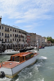 A wooden motorboat navigates a canal lined with historic buildings featuring multiple stories and balconies. Gondolas are moored along the waterway with pedestrians walking on the adjacent promenade. The sky is partly cloudy, adding a serene backdrop to the vibrant scene.