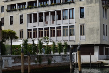 A multi-story building with numerous windows and a balcony decorated with plants. Below there is a garden area with leafy arches and an outdoor seating area, partially shaded by umbrellas. Wooden posts stand in front of the garden, with birds perched on them.