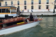 A motorboat with a wooden finish glides through a canal. A man stands at the helm, steering the boat past a dock labeled 'Ferrovia'. In the background, a sidewalk runs alongside a beige and pink building, where several people walk by, some pulling luggage on wheels.