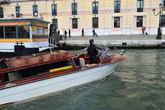 A motorboat with a wooden finish glides through a canal. A man stands at the helm, steering the boat past a dock labeled 'Ferrovia'. In the background, a sidewalk runs alongside a beige and pink building, where several people walk by, some pulling luggage on wheels.