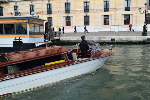 A motorboat with a wooden finish glides through a canal. A man stands at the helm, steering the boat past a dock labeled 'Ferrovia'. In the background, a sidewalk runs alongside a beige and pink building, where several people walk by, some pulling luggage on wheels.