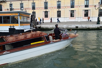 A motorboat with a wooden finish glides through a canal. A man stands at the helm, steering the boat past a dock labeled 'Ferrovia'. In the background, a sidewalk runs alongside a beige and pink building, where several people walk by, some pulling luggage on wheels.