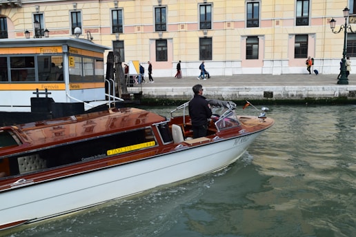 A motorboat with a wooden finish glides through a canal. A man stands at the helm, steering the boat past a dock labeled 'Ferrovia'. In the background, a sidewalk runs alongside a beige and pink building, where several people walk by, some pulling luggage on wheels.
