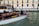 A motorboat with a wooden finish glides through a canal. A man stands at the helm, steering the boat past a dock labeled 'Ferrovia'. In the background, a sidewalk runs alongside a beige and pink building, where several people walk by, some pulling luggage on wheels.
