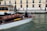 A motorboat with a wooden finish glides through a canal. A man stands at the helm, steering the boat past a dock labeled 'Ferrovia'. In the background, a sidewalk runs alongside a beige and pink building, where several people walk by, some pulling luggage on wheels.