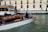 A motorboat with a wooden finish glides through a canal. A man stands at the helm, steering the boat past a dock labeled 'Ferrovia'. In the background, a sidewalk runs alongside a beige and pink building, where several people walk by, some pulling luggage on wheels.