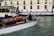 A motorboat with a wooden finish glides through a canal. A man stands at the helm, steering the boat past a dock labeled 'Ferrovia'. In the background, a sidewalk runs alongside a beige and pink building, where several people walk by, some pulling luggage on wheels.