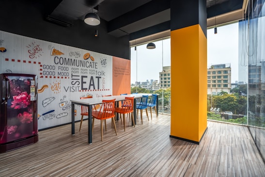 A modern office space with large windows offering a city view, featuring a colorful interior design with brightly painted walls and an accent wall with playful food-related words and illustrations. The room has a long table surrounded by orange and blue chairs, and a refrigerator with a floral design is positioned against the wall.