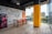 A modern office space with large windows offering a city view, featuring a colorful interior design with brightly painted walls and an accent wall with playful food-related words and illustrations. The room has a long table surrounded by orange and blue chairs, and a refrigerator with a floral design is positioned against the wall.