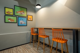 A modern dining area with three orange high stools arranged neatly under a long wooden counter. The walls are adorned with colorful framed art showcasing various food items like samosa, dosa, and chai. A single pendant light provides illumination from above, casting subtle shadows on the light gray walls. The floor is covered with wooden planks, adding a warm touch to the simple and clean design.