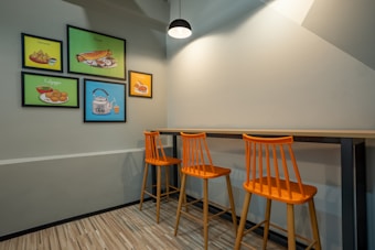 A modern dining area with three orange high stools arranged neatly under a long wooden counter. The walls are adorned with colorful framed art showcasing various food items like samosa, dosa, and chai. A single pendant light provides illumination from above, casting subtle shadows on the light gray walls. The floor is covered with wooden planks, adding a warm touch to the simple and clean design.