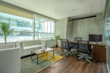 A modern office space with a clean, minimalist design featuring a large window allowing for natural light. There is a white couch with a wooden coffee table in front of it, decorated with a small white vase. The office also includes a wooden desk with two office chairs and a computer monitor. Potted plants are placed around the space, adding greenery to the environment. The floor is wooden, complementing the overall elegance of the room.