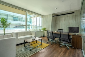 A modern office space with a clean, minimalist design featuring a large window allowing for natural light. There is a white couch with a wooden coffee table in front of it, decorated with a small white vase. The office also includes a wooden desk with two office chairs and a computer monitor. Potted plants are placed around the space, adding greenery to the environment. The floor is wooden, complementing the overall elegance of the room.