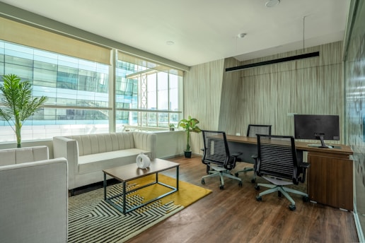 A modern office space with a clean, minimalist design featuring a large window allowing for natural light. There is a white couch with a wooden coffee table in front of it, decorated with a small white vase. The office also includes a wooden desk with two office chairs and a computer monitor. Potted plants are placed around the space, adding greenery to the environment. The floor is wooden, complementing the overall elegance of the room.