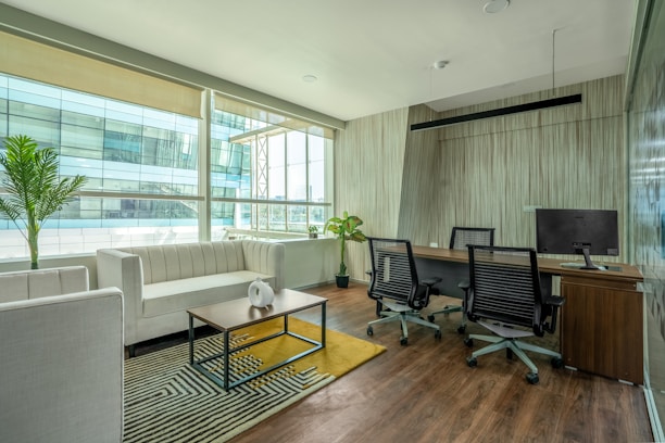 A modern office space with a clean, minimalist design featuring a large window allowing for natural light. There is a white couch with a wooden coffee table in front of it, decorated with a small white vase. The office also includes a wooden desk with two office chairs and a computer monitor. Potted plants are placed around the space, adding greenery to the environment. The floor is wooden, complementing the overall elegance of the room.