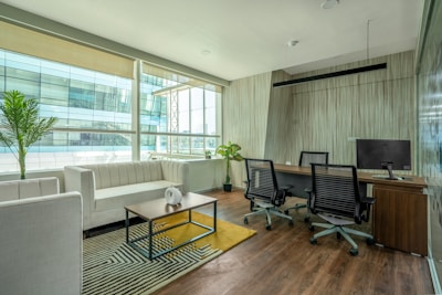 A modern office space with a clean, minimalist design featuring a large window allowing for natural light. There is a white couch with a wooden coffee table in front of it, decorated with a small white vase. The office also includes a wooden desk with two office chairs and a computer monitor. Potted plants are placed around the space, adding greenery to the environment. The floor is wooden, complementing the overall elegance of the room.