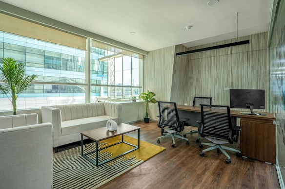 A modern office space with a clean, minimalist design featuring a large window allowing for natural light. There is a white couch with a wooden coffee table in front of it, decorated with a small white vase. The office also includes a wooden desk with two office chairs and a computer monitor. Potted plants are placed around the space, adding greenery to the environment. The floor is wooden, complementing the overall elegance of the room.