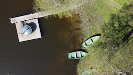 An aerial view of a small dock on the edge of a body of water. There is a circular structure on the dock, possibly a hot tub or similar feature. Two small green boats are moored near the dock, one of which is partly on the grassy shore. The area around the water has green vegetation and trees with partial shadows visible. The water is dark, indicating depth, or it may be due to the reflection of the sky.