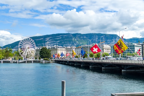 A picturesque cityscape features a large ferris wheel on the left next to a body of water. Multiple flags, including those of Switzerland, are displayed on a bridge across from the water. In the background, there is a range of lush green hills under a partly cloudy sky.