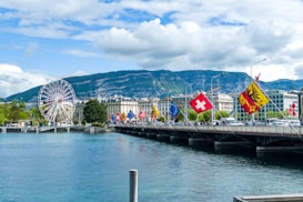 A picturesque cityscape features a large ferris wheel on the left next to a body of water. Multiple flags, including those of Switzerland, are displayed on a bridge across from the water. In the background, there is a range of lush green hills under a partly cloudy sky.
