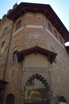 A historic stone building with a multi-tiered facade, featuring arched windows and decorative elements. The architectural style is traditional, with intricate carvings and a painted panel on the wall depicting historical figures.