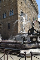 A grand stone fountain features a large white marble statue surrounded by several smaller bronzed figures. Water cascades from the centerpiece, adding a dynamic element. The fountain is set against the backdrop of an ornate, historic stone building with arched windows and flags, suggesting a European architectural style.