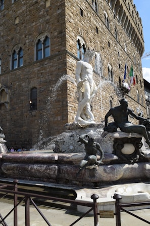A grand stone fountain features a large white marble statue surrounded by several smaller bronzed figures. Water cascades from the centerpiece, adding a dynamic element. The fountain is set against the backdrop of an ornate, historic stone building with arched windows and flags, suggesting a European architectural style.