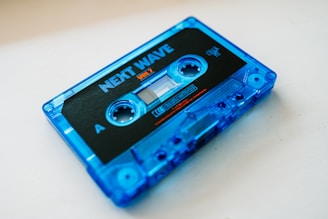 A vintage blue cassette tape with the label 'NEXT WAVE VOL 7' is placed diagonally on a light surface. The cassette features a black and blue color scheme with visible text and a clear window for viewing the tape.
