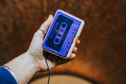 A hand is holding a portable cassette player with a visible cassette inside labeled 'Next Wave'. The player has a mix of purple and white with a yellow button.