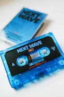 A blue audio cassette tape with the label 'Next Wave Vol 7' is prominently displayed. The cassette is partially transparent, showcasing the internal spools and tape. In the background, a matching blue cassette case with the same 'Next Wave' design is visible, slightly out of focus.