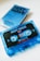 A blue audio cassette tape with the label 'Next Wave Vol 7' is prominently displayed. The cassette is partially transparent, showcasing the internal spools and tape. In the background, a matching blue cassette case with the same 'Next Wave' design is visible, slightly out of focus.