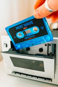 A close-up view of a hand holding a blue cassette tape labeled 'Next Wave Vol 1.' The cassette tape is being inserted into a silver cassette player, which has its front door open.