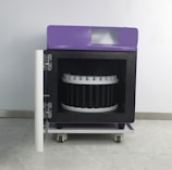 A wheeled laboratory or industrial machine with a purple top and a front door open to reveal a circular arrangement inside, possibly for holding samples or items. It is set against a plain wall.