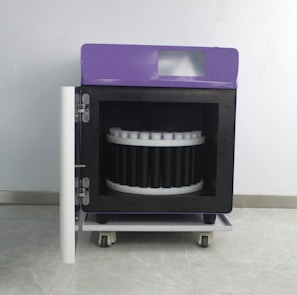 A wheeled laboratory or industrial machine with a purple top and a front door open to reveal a circular arrangement inside, possibly for holding samples or items. It is set against a plain wall.