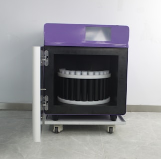 A wheeled laboratory or industrial machine with a purple top and a front door open to reveal a circular arrangement inside, possibly for holding samples or items. It is set against a plain wall.