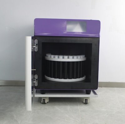 A wheeled laboratory or industrial machine with a purple top and a front door open to reveal a circular arrangement inside, possibly for holding samples or items. It is set against a plain wall.