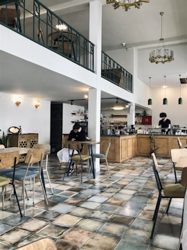 A spacious cafe with a modern and minimalist design features wooden furniture and tiled floors. There is a mezzanine level with a railing, adding to the open feel of the space. A person is sitting at a table using a phone, and there is another person working behind the counter with various coffee-making equipment. Pendant lights hang from the ceiling, and decorative elements such as plants and wall fixtures add to the ambiance.
