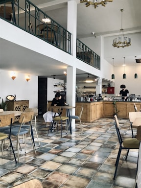 A spacious cafe with a modern and minimalist design features wooden furniture and tiled floors. There is a mezzanine level with a railing, adding to the open feel of the space. A person is sitting at a table using a phone, and there is another person working behind the counter with various coffee-making equipment. Pendant lights hang from the ceiling, and decorative elements such as plants and wall fixtures add to the ambiance.