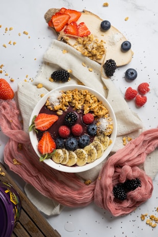 A vibrant breakfast setup featuring a bowl filled with an acai smoothie topped with fresh fruit such as strawberries, blueberries, raspberries, blackberries, and banana slices. The bowl also includes granola and seeds as garnish. Surrounding the bowl are scattered granola, whole berries, and a sliced apple topped with strawberries and granola. The arrangement is enhanced by soft fabric and a wooden surface.