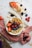A vibrant breakfast setup featuring a bowl filled with an acai smoothie topped with fresh fruit such as strawberries, blueberries, raspberries, blackberries, and banana slices. The bowl also includes granola and seeds as garnish. Surrounding the bowl are scattered granola, whole berries, and a sliced apple topped with strawberries and granola. The arrangement is enhanced by soft fabric and a wooden surface.
