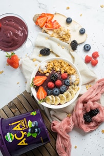 A well-arranged flat lay of a vibrant acai bowl garnished with banana slices, strawberries, blueberries, blackberries, granola, and chia seeds, set on a white surface with a fabric napkin. Next to the bowl is a glass of acai smoothie, some loose fruits like raspberries and blackberries, a slice of toast topped with strawberries and granola, and a container of acai puree. The overall setting includes wooden planks and a pink cloth for visual contrast.