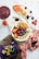 A well-arranged flat lay of a vibrant acai bowl garnished with banana slices, strawberries, blueberries, blackberries, granola, and chia seeds, set on a white surface with a fabric napkin. Next to the bowl is a glass of acai smoothie, some loose fruits like raspberries and blackberries, a slice of toast topped with strawberries and granola, and a container of acai puree. The overall setting includes wooden planks and a pink cloth for visual contrast.