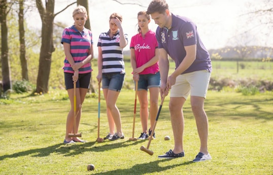 Four young adults are engaging in a game of croquet on a grassy field. They are dressed in casual, sporty attire with striped polo shirts. The background features trees and an open field, suggesting a pleasant, sunny day.