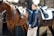 A woman is standing between two saddled horses, holding onto their reins. She wears a blue jacket with the word 'TEAM' visible on the sleeve. The horses are a rich brown color, and saddles are clearly in view. The background shows a stable setting with brick walls.