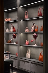 A display of luxury handbags arranged neatly on shelves inside a well-lit retail store. The bags vary in color from pink and orange to white and brown. Some mannequins wear accessories like sunglasses and scarves alongside the handbags.