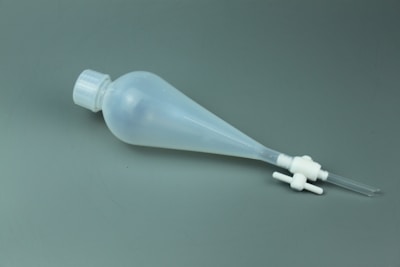 A translucent plastic separatory funnel with a conical shape lying on a grey surface. It has a screw cap on the top, a stopcock valve mechanism at the bottom, and a small nozzle extending from the valve.