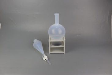 Two laboratory glassware items are placed against a neutral gray background. A round-bottomed flask sits on a small white stand, while a separatory funnel lies on its side nearby.